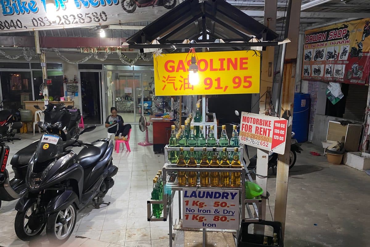 A roadside stall in Phuket selling Gasoline 91/95 in glass bottles, next to a 'Motorbike for Rent' sign